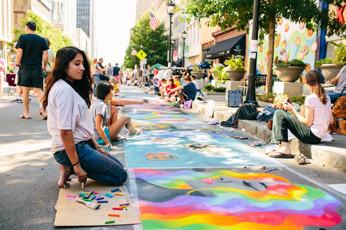 Young people sitting on the street in front of paintings.